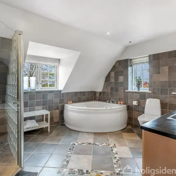 Badeværelse med rundt badekar i hjørnet, placeret ved vinduer med udsigt til træer. Bruseren er til venstre, og der er en håndvask med sort bordplade til højre. Flisegulvet har stenudsmykning.