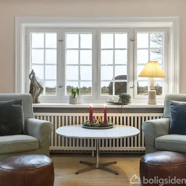 To lænestole med puder står overfor hinanden ved et rundt bord med røde stearinlys. En lampe og dekorationer er placeret på vindueskarmen i et lyst, hyggeligt rum.