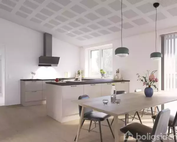 Et moderne køkken med sandfarvede skabe og en central ø, der har kogeplade og emhætte. To hængelamper over et spisebord med stole. Store vinduer lader naturligt lys ind.