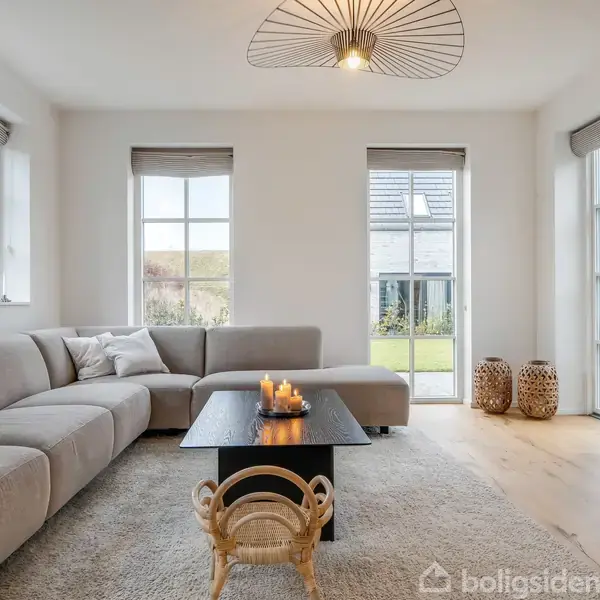 En lys stue med en stor grå sofa sidder omkring et sort sofabord, hvorpå der står tændte lys. Rummet har store vinduer og moderne indretning.
