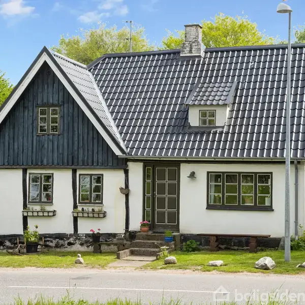 Et hyggeligt, hvidmalet hus med sort tag ligger ved en stille vej. Huset har blomster i vindueskasser under vinduerne. En grøn have omgiver bygningen med frodige træer i baggrunden.