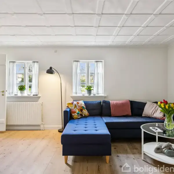 Mørkeblå hjørnesofa med farverige puder står i en lys stue. Rundt sofabord med blomster, skriveøvelse hænger på væggen, og to vinduer slipper dagslys ind.