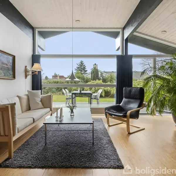 En lys stue har en sofa og en lænestol ved et sofabord med planter. Store vinduer viser en have med træer og udendørs møbler. En stor plante står til højre.