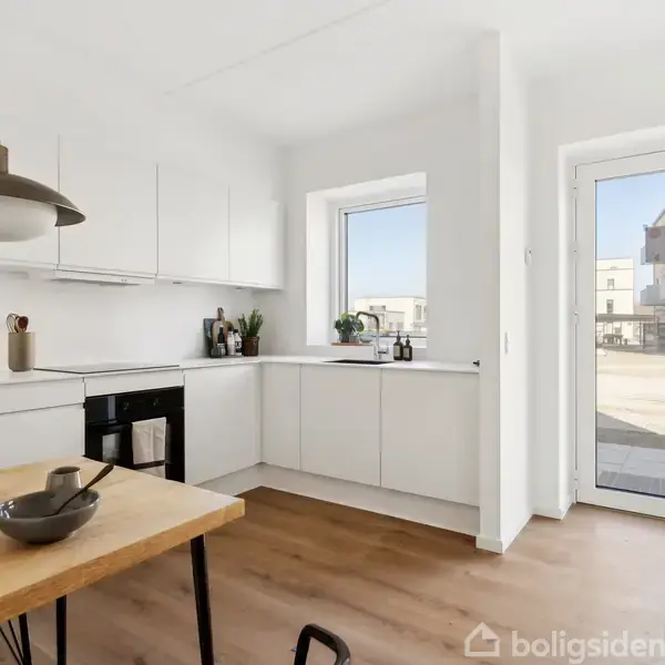 Moderne køkken med hvide skabe og sort ovn. Spisebord med skål og klud. Stor glasdør og vindue giver lys. Bygninger udenfor ses gennem døren. Ingen tekst i billedet.