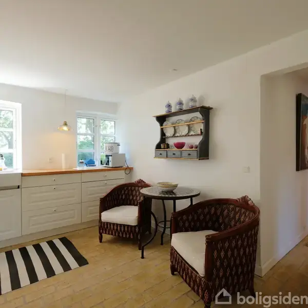 Et køkken med to kurvestole og et rundt bord. Køkkenet har hvide skabe, et sort-hvidt tæppe, og hylder med porcelæn. Lys kommer fra to vinduer bag vasken.