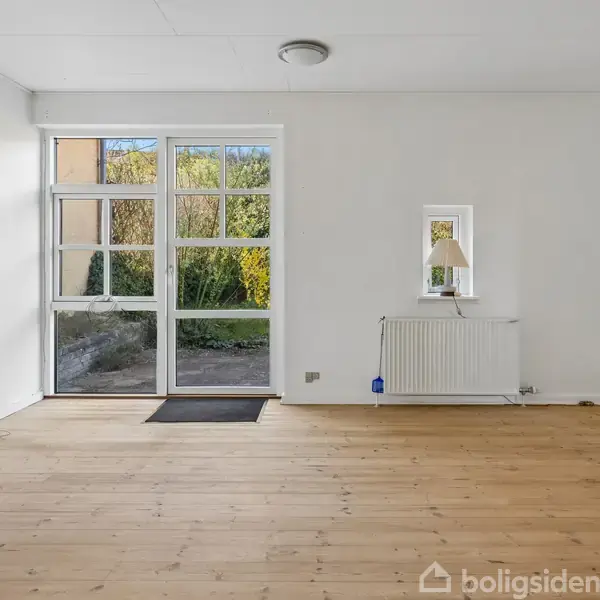 Stue med trægulv og store vinduer, der fører til en have. En lille radiator med lampe ved siden af, hvide vægge.