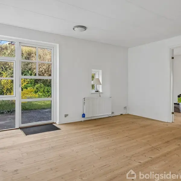Stue med trægulv, store vinduer og havedør, der åbner ud mod en frodig have. Et radiator og lysarmatur ses, samt adgang til et tilstødende rum med flyttekasser.