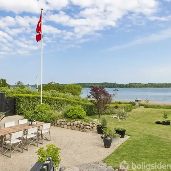 Et udendørs spiseområde med et bord og stole står på en terrasse ved siden af en flagstang med et flag i vinden. I baggrunden ses en sø og frodig natur.