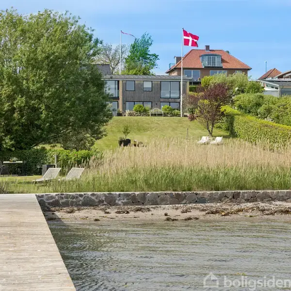 Et velholdt hus med modernistisk design står på en grøn, skrånende grund. En dansk flagstang er til stede. Foran er en træbro, der strækker sig ud i en rolig sø.