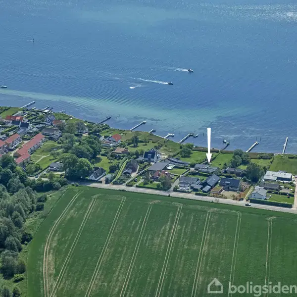 Kystområde med huse langs en vej og marker. En stor, skovklædt park ligger ved siden af kystlinjen. Flere både ses på det blå hav.