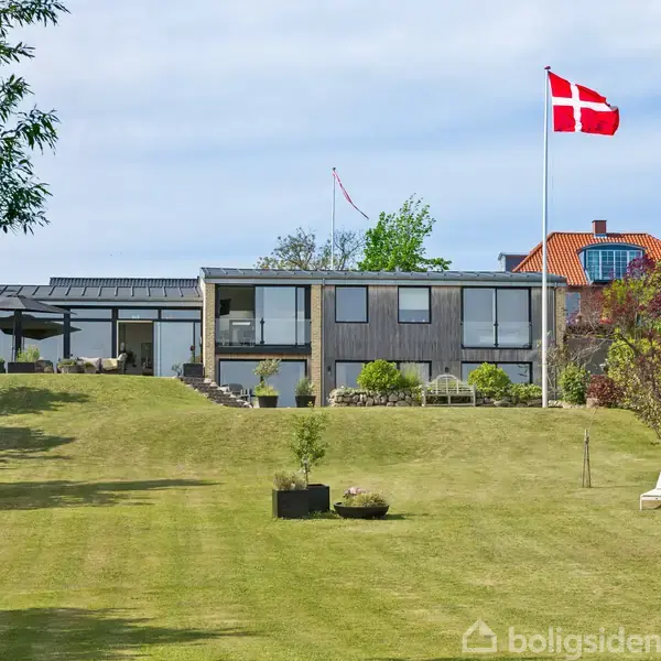 En moderne villa med store vinduer står på en velplejet græsplæne. En dansk flagstang med dannebrog til højre, omgivet af buske, træer og to liggestole på plænen.