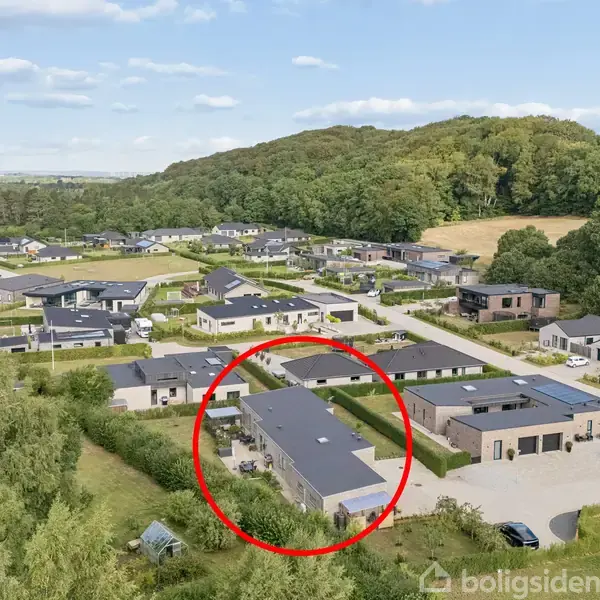Et moderne boligområde med mange huse er omgivet af skovklædte bakker. Et specifikt hus er indrammet med en rød cirkel, hvilket fremhæver det blandt de andre hjem og grønne områder.