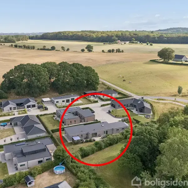 Et hus omgivet af en have er markeret med en rød cirkel. Det ligger i et boligområde tæt ved åbne marker og skovbevoksning.