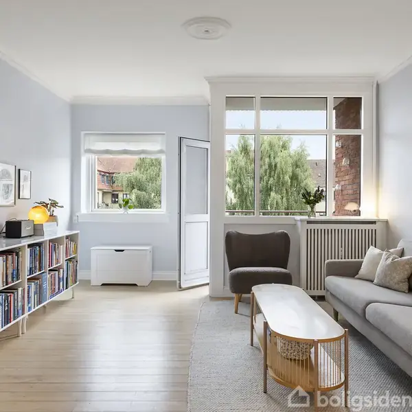 Stue med sofa, sofabord og lænestol ved siden af store vinduer. Bogreol fyldt med bøger og væghængt TV. Lyse vægge og trægulv skaber en lys og indbydende atmosfære.