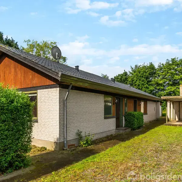 Et murstenshus med sort tag står ved siden af en carport. Huset er omgivet af grøn hæk og træer, under en delvist overskyet himmel.