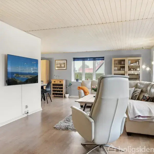 Stue med moderne lænestol i centrum, vendt mod en vægmonteret fladskærm. Omgivet af sofaer, et tæppe, og reol, mens store vinduer giver lysindfald.