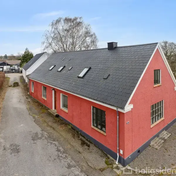 Rødt hus med gråt tag står stille ved en vej i et boligkvarter. En lille grusvej løber langs siden, omgivet af hække og træer i baggrunden.