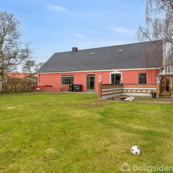 Et rødt hus med sort tag står i en grøn have. En bold ligger på græsplænen, omgivet af træer og en hæk.