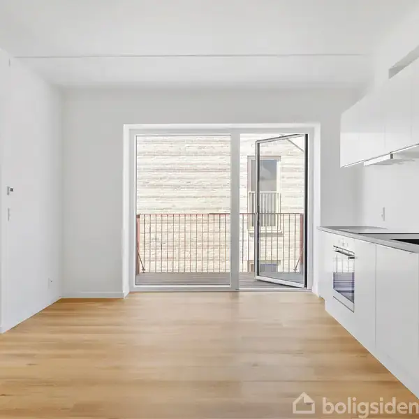 Et lyst køkkenrum med hvide skabe og integreret ovn i højre side. En åben altandør fører til en lille balkon. Trægulve skaber en moderne atmosfære.
