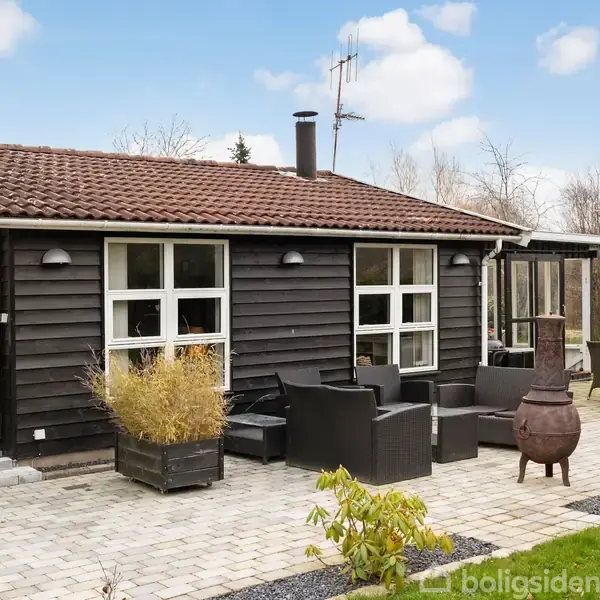 Et sortmalet træhus med rødt tegltag står på en flisebelagt terrasse. Havemøbler og en kamin er placeret udenfor. Baggrunden viser en have med buske og træer.
