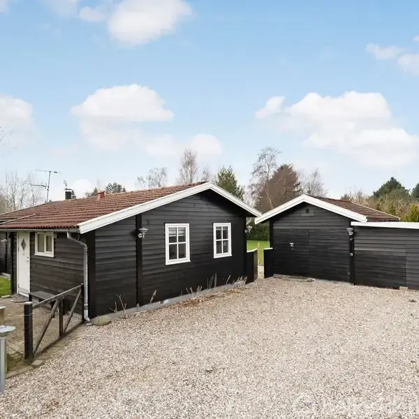 Sort træhus med rødt tag står på en grusbelagt indkørsel. Ved siden af huset er en garage. Omkring huset er der grønt græs og træer i baggrunden under en blå himmel med få skyer.