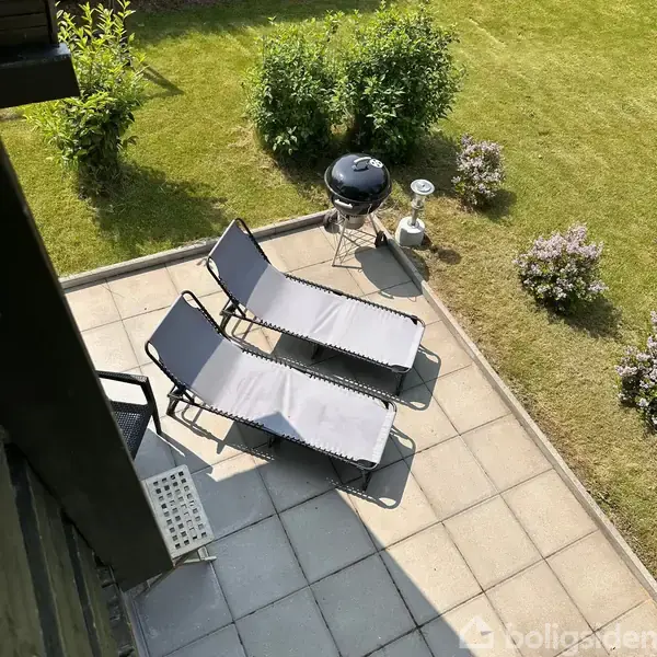 To liggestole står på en flisebelagt terrasse ved siden af en kuglegrill og en lanterne. Der er græsplæne og buske i baggrunden.