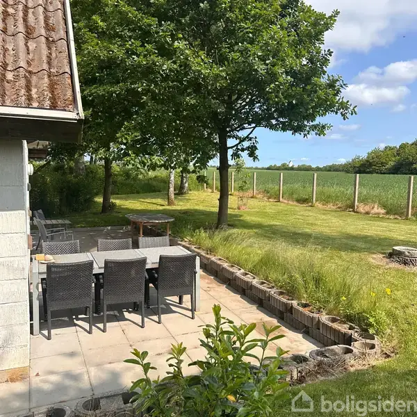 Terrassemøbler med otte stole står på flisebelagt terrasse, omgivet af en have med græsplæne og træer. I baggrunden ses marker under en blå himmel med skyer.