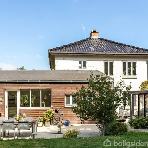 To-etagers hus med en træbeklædt tilbygning og sort tag. I forgrunden ses en terrasse med et spisebord, stole og grill. Haven er frodig med græs og buske.