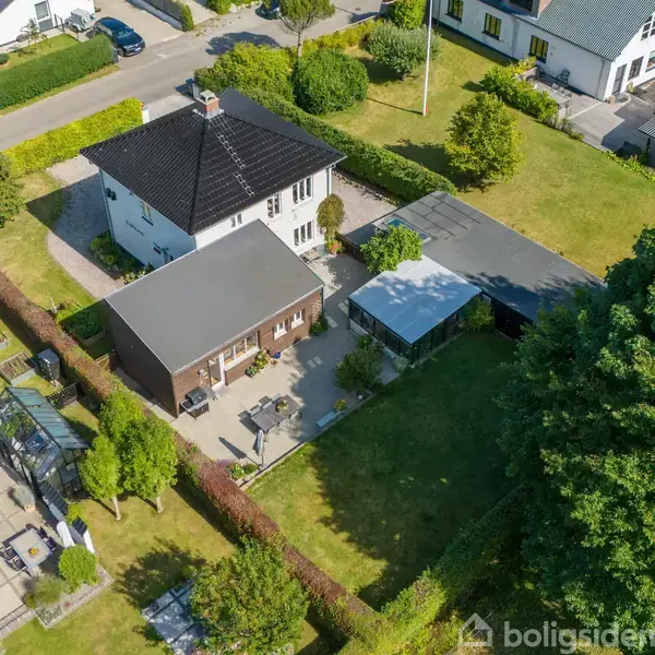 Et hus med sort tag står i en velplejet have med terrasse og garage. Omgivet af andre huse, træer og græsplæner, danner det en del af en rolig beboelsesvej.