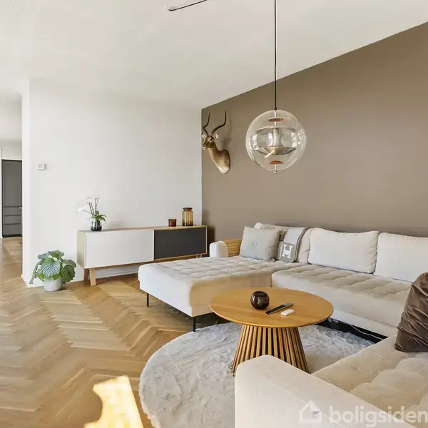 En moderne stue med en stor, beige hjørnesofa, står ved en brun væg med et hjortehoved. Et rundt sofabord er foran sofaen. Trægulv strækker sig mod en lys gang.