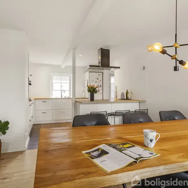 En træbord er dækket med en magasin og en kop, omgivet af moderne stole i et rummeligt, lyst køkken-alrum med hvide skabe, grønne planter og en unik sort-hængende lampe.