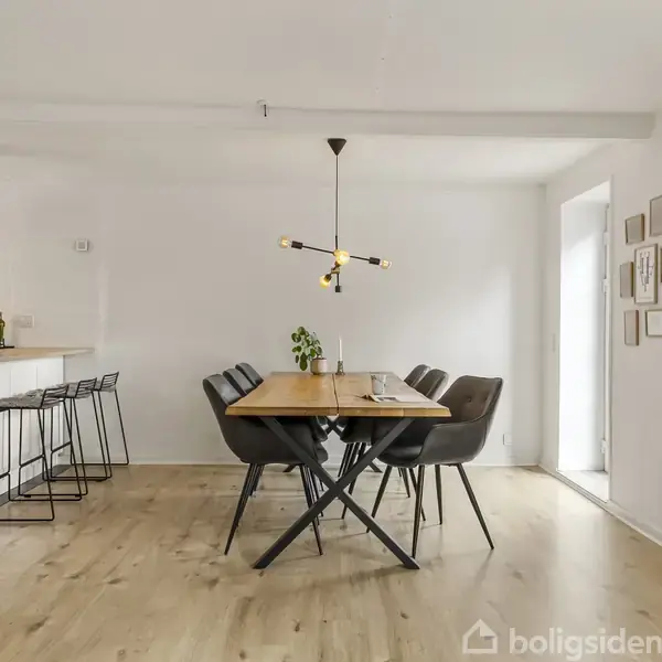 Et spisebord med seks stole står i en lys spisestue. Bordet har en moderne pendel hængende over det. I baggrunden ses et køkken med barstole og blomster.
