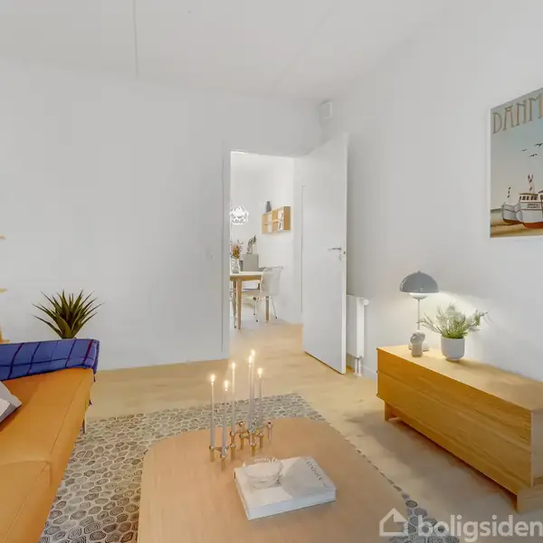 En stue med en orange sofa dekoreret med puder, et sofabord med lys og bøger, og en plakat med teksten "DANMARK". En åbning fører til et andet rum med spisebord.