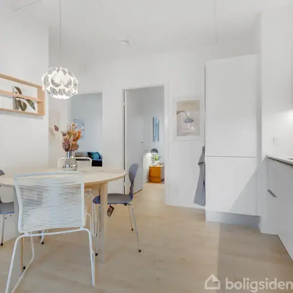 Rundt spisebord med fire stole og vase med blomster i en moderne, lys spisestue; hvidt køkken med indbyggede skabe til højre, dekoreret med en elegant lampe.