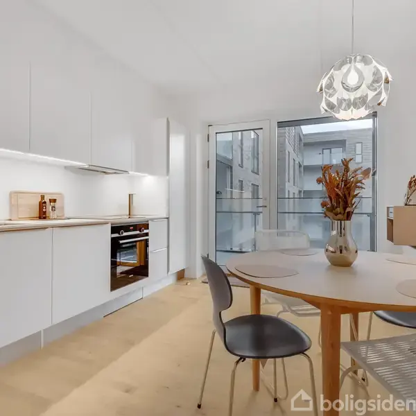 Et moderne køkken med hvide skabe og en indbygningsovn. En rund træbord står i centrum med stole omkring. Store vinduer i baggrunden giver lys.