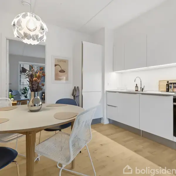 Rundt spisebord med stole står i et lyst moderne køkken med hvidt inventar og en indbygget ovn. En lampe hænger over bordet, og en åben dør fører til et andet rum.