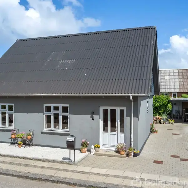 Gråt hus med sort tag står på en solrig gade. Foran huset er der blomsterkrukker og en postkasse. Til højre er en indkørsel med fliser og et skur.