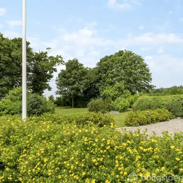 Flagstang står i stille have omgivet af tætte buske med gule blomster og frodig græsplæne; baggrund viser store træer under en let skyet, blå himmel.