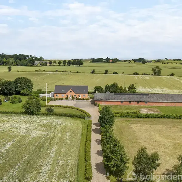 En landejendom ligger mellem marker, med en lang grusvej, grønne træer og buske omkring. Bygningerne inkluderer et rødt hus med sort tag og en tilstødende lade.