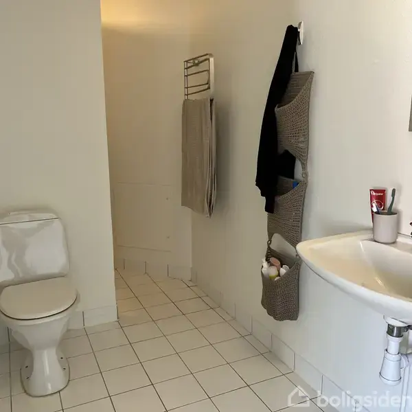 Toilet står ved siden af vask med tandkrus og sæbe. Badeværelse med lyse fliser og vægmonteret opbevaring i hjørnet. Håndklæder hænger på stang ved bruseniche.