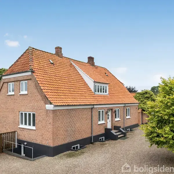 En rød murstensvilla med rødt tegltag står på en grusvej omgivet af grønne træer. Huset har flere vinduer og en lille trappe ved hoveddøren.