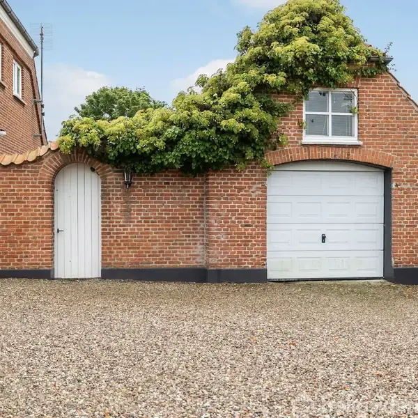 Garagebygning med rød murstensfacade og hvid port, tilgroet med grønne planter. Gruset gårdsplads omgiver bygningen, og en buet dør findes til venstre.