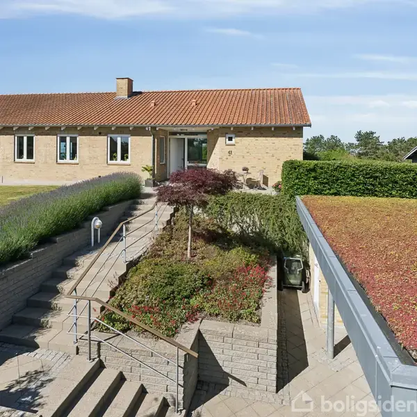 Et gult murstenshus med rødt tegltag står på en velplejet grund, omgivet af grønne buske og blomster. En trappe leder op til hovedindgangen, og himlen er klar og blå.