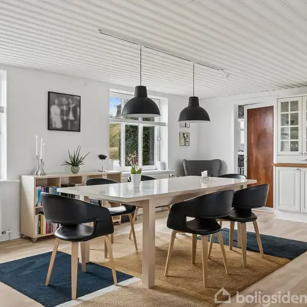 Spisebord med seks sorte stole står i en lys spisestue med træbjælkeloft og to store vinduer. To sorte lamper hænger over bordet. Skabssektion og planter skaber hyggelig atmosfære.