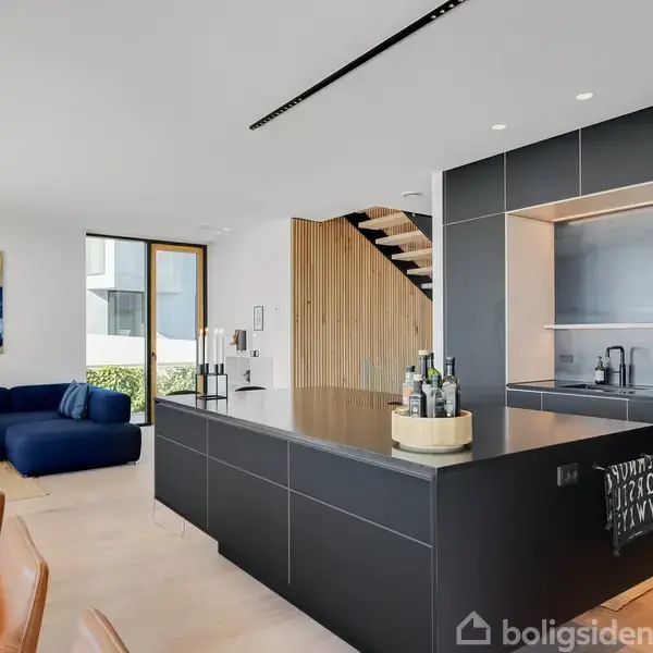 Køkken-ø står centralt med flasker ovenpå; moderne køkkenindretning i en lys stue med spisebord, blå sofa, trappe og stort vindue. Maleri hænger på væggen.