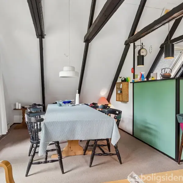 Et spisebord er dækket med en blå dug og omgivet af sorte stole. Rummet har skrå loftsbjælker, grønne skillevægge, og et stort vindue med hvide gardiner.