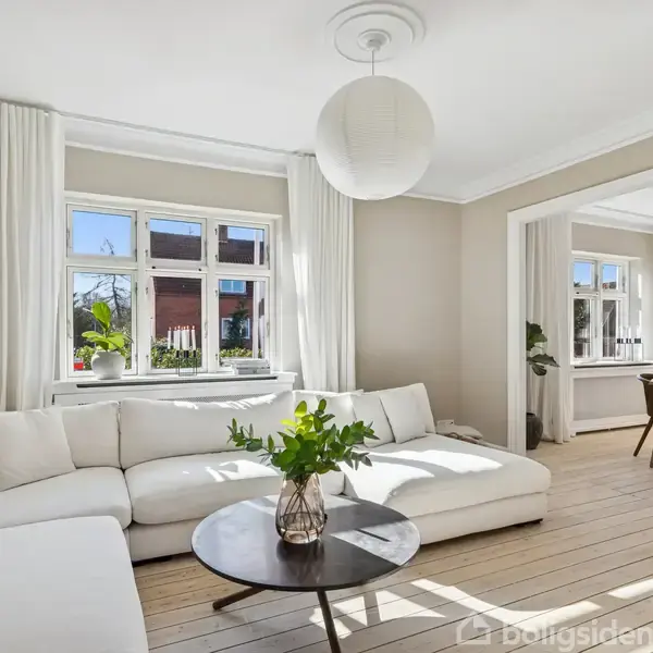 Hvid sofa står i stue med lyse vægge. Rundt sofabord har planter i vaser. Store vinduer lukker dagslys ind. Åbning fører til spisestue med træbord og stole.