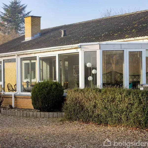 Et enfamiliehus med en stor glasveranda står i en grusbelagt have. Buske omkranser huset, der har en synlig skorsten og gult murværk.