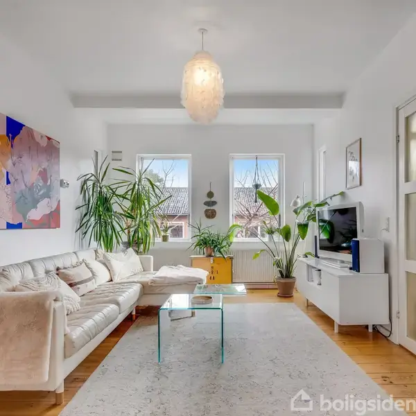 En beige sofa står mod en væg med en farverig plakat, omgivet af grønne planter. Rummet har store vinduer, udsigt til tage, og en gul kiste fungerer som bord.