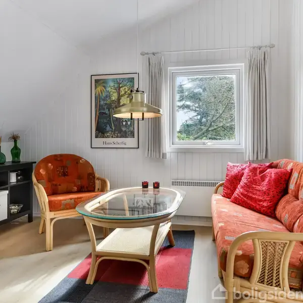 En stue med en glasbord omgivet af to flettede sofaer med orange puder. En plakat med teksten "Henrik Sohlberg" hænger på væggen. Et vindue viser træer udenfor.